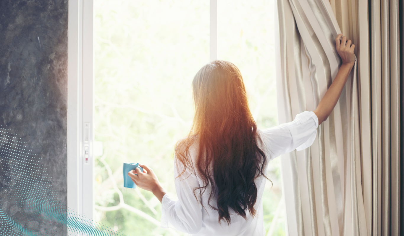 A person looking out of window shining through sunlight whilst holding back the curtain 