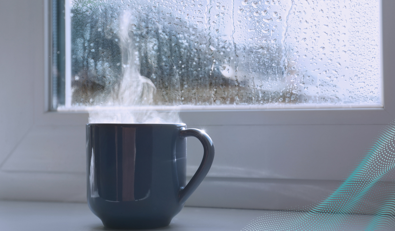 A mug is sat on a window sill steaming, whilst rain is pouring outside