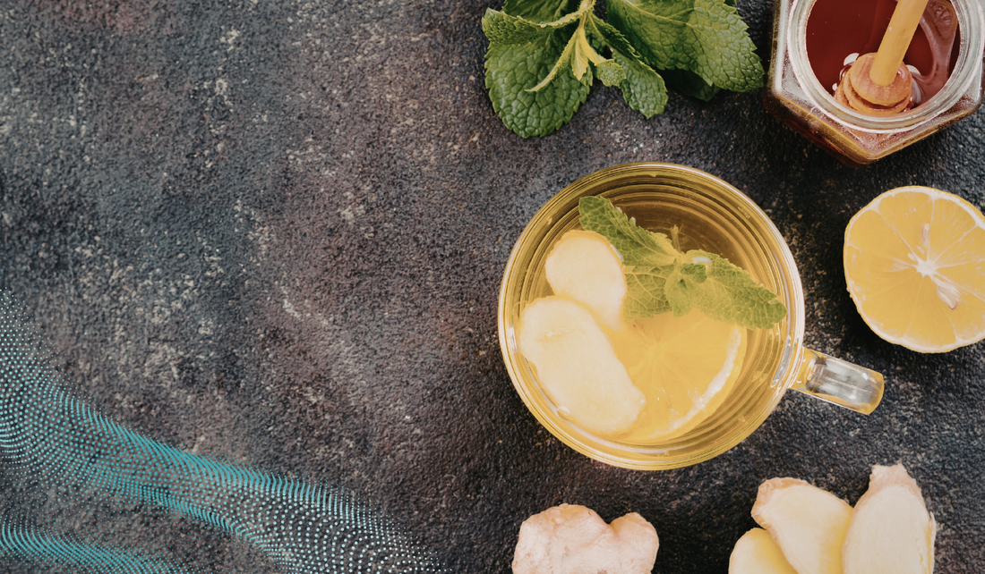 A glass mug of lemon and ginger tea garnished with fresh mint, surrounded by sliced lemon, ginger pieces, mint leaves, and a jar of honey