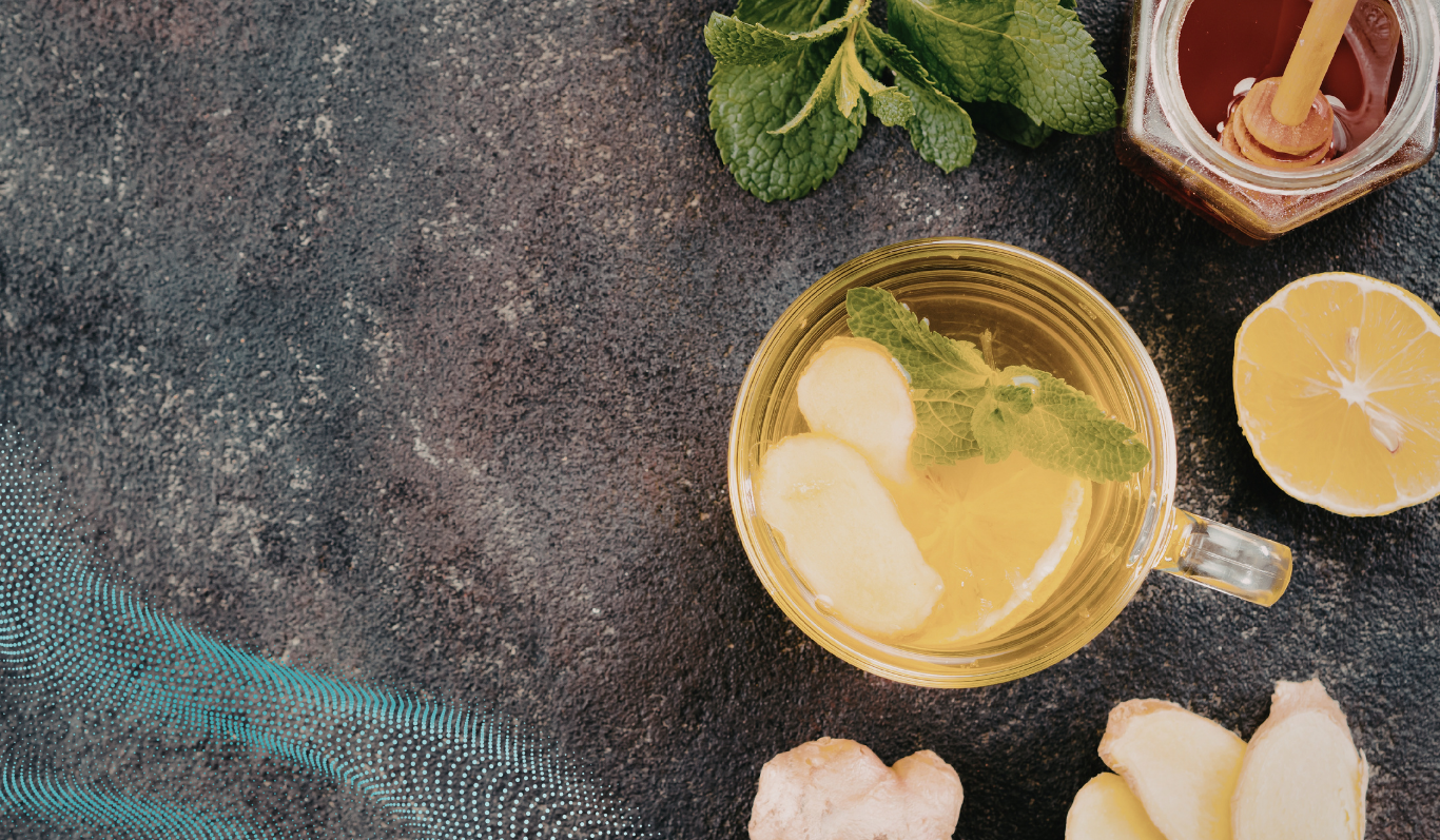 A glass mug of lemon and ginger tea garnished with fresh mint, surrounded by sliced lemon, ginger pieces, mint leaves, and a jar of honey
