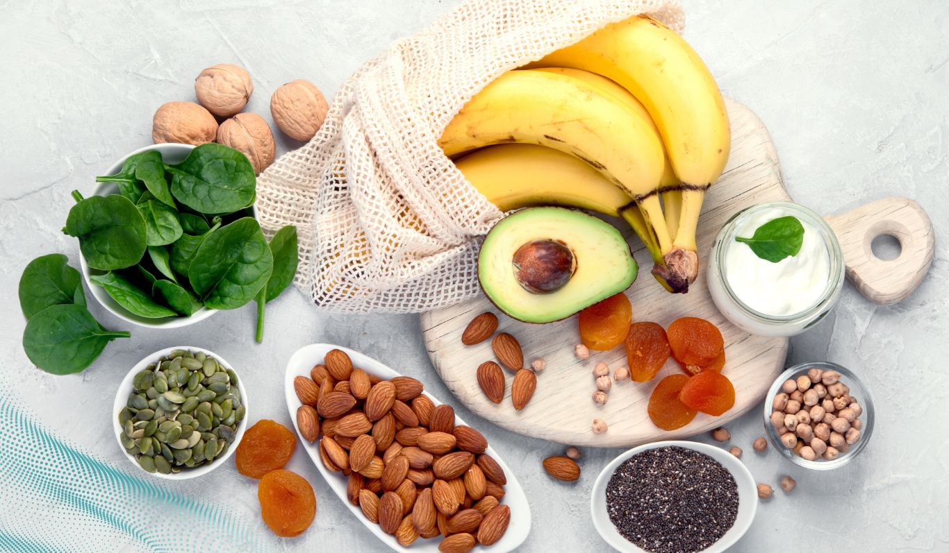 A lay of bananas, avocado, spinach, nuts, seeds, dried apricots, chickpeas, and yogurt on a light surface.